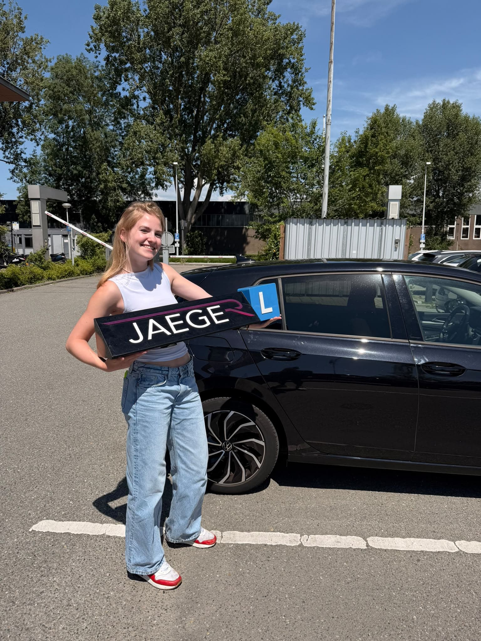 Geslaagde leerling met een Jaeger-bord naast de lesauto op een zonnige dag